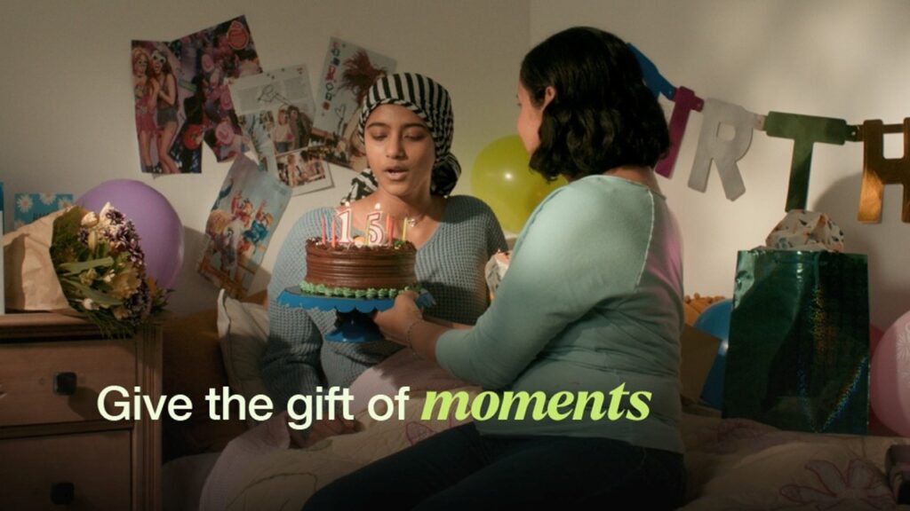 Mother and daughter on a bed with a birthday cake, with overlay text reading 'give the gift of moments'