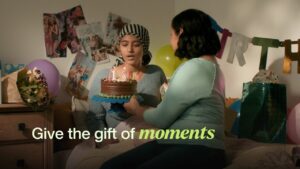 Mother and daughter on a bed with a birthday cake, with overlay text reading 'give the gift of moments'